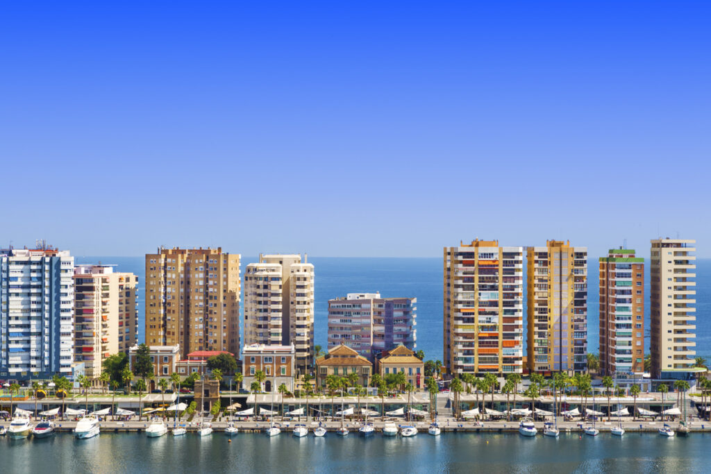 Vista panorámica del puerto de Málaga con modernos edificios residenciales frente al mar, barcos atracados en el muelle y un cielo azul despejado, destacando el atractivo del mercado inmobiliario en la zona costera.